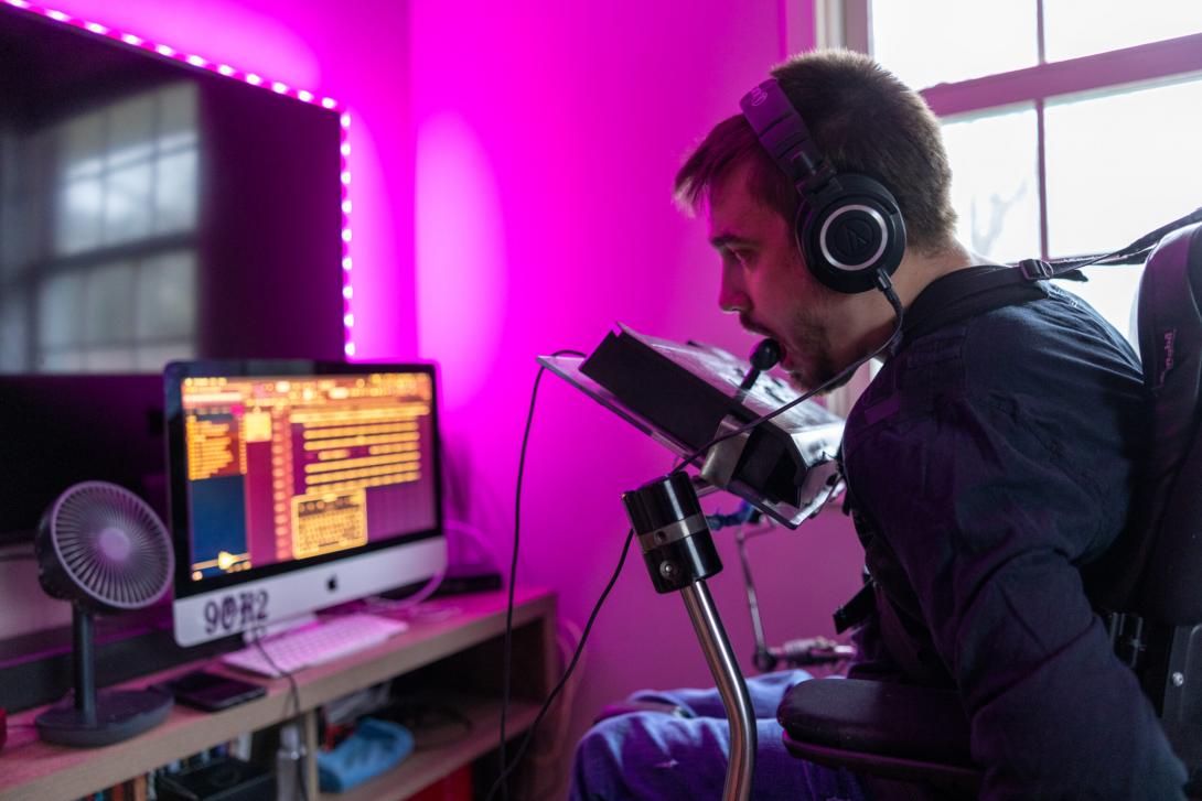 1451 - A person wearing over-the-ear headphones leans forward to use a mouth joystick controller on a raised device attached to the front to their power wheelchair while looking at a computer monitor in front of them. 