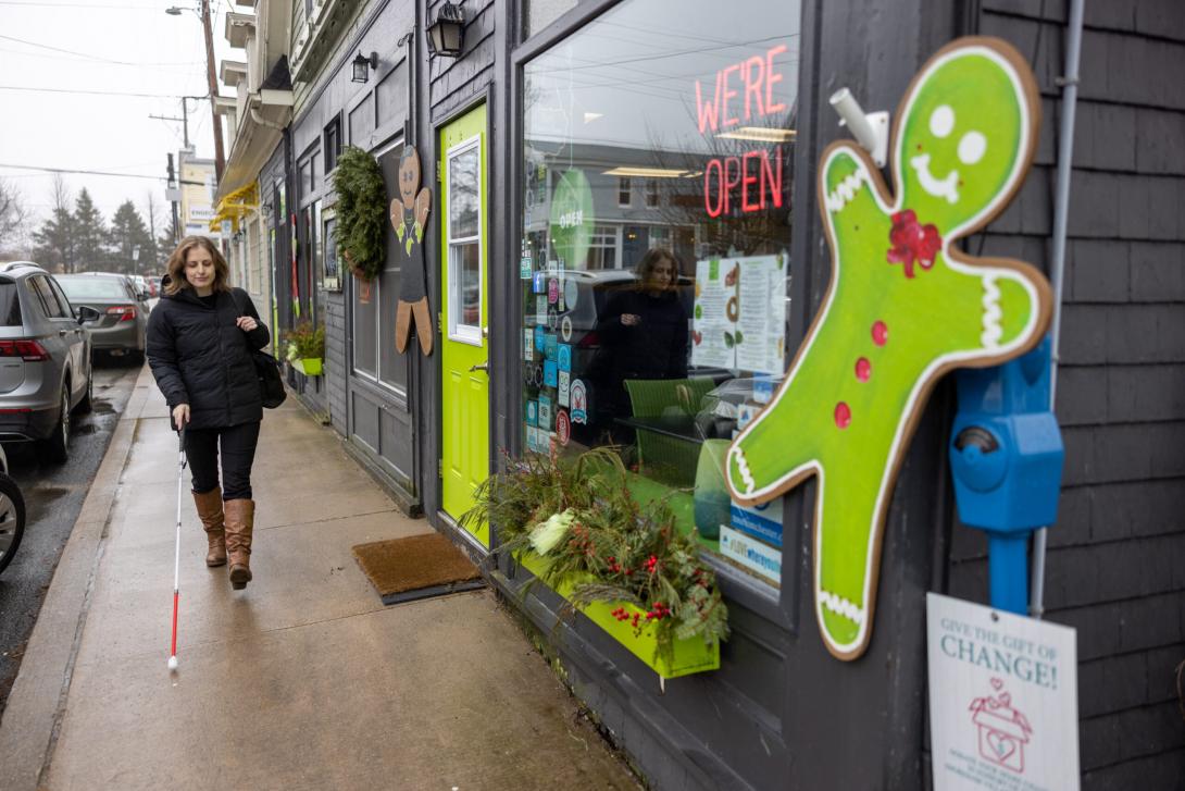 6497 A light skinned woman navigates a sidewalk between storefronts and parked cars in a town centre using a white cane. She is moving past a restaurant decorated with gingerbread people.  