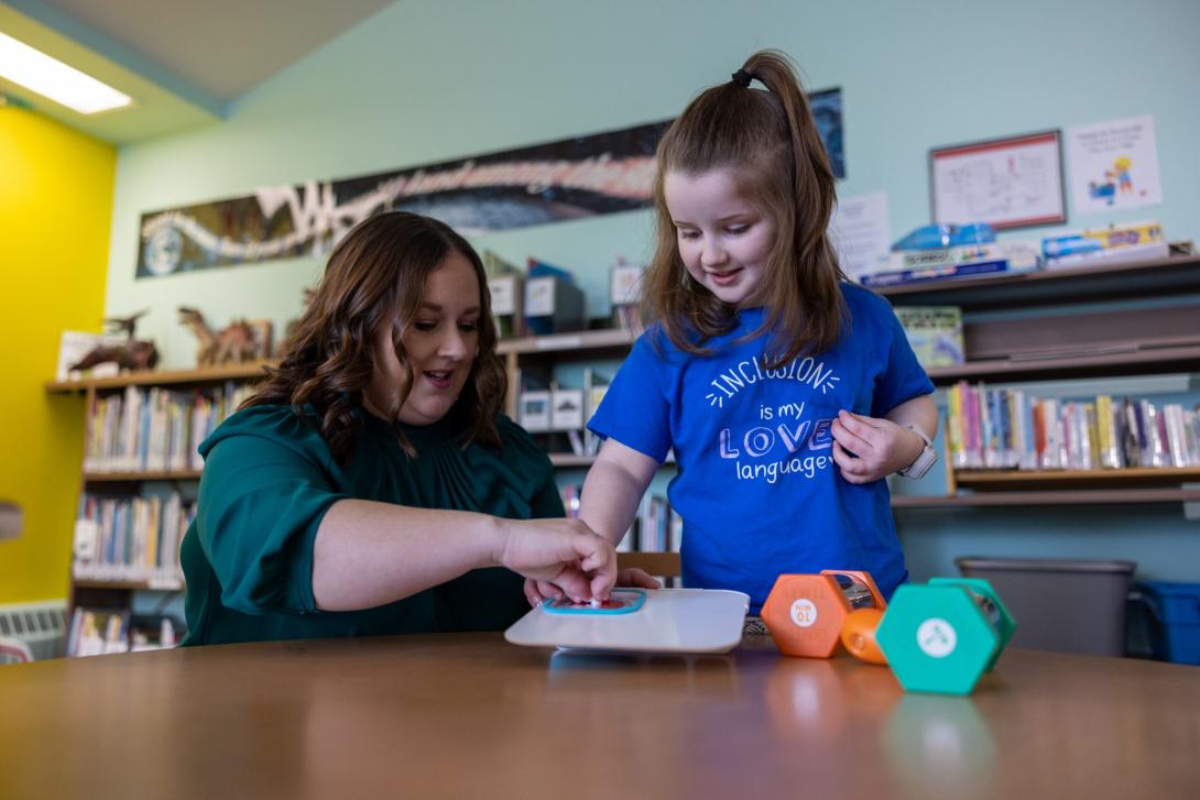 7439 - A light skinned young child with long hair and an adult beside them explore visual time telling tools on a table in a library. The child is wearing a t-shirt that says “inclusion is my love language.”  