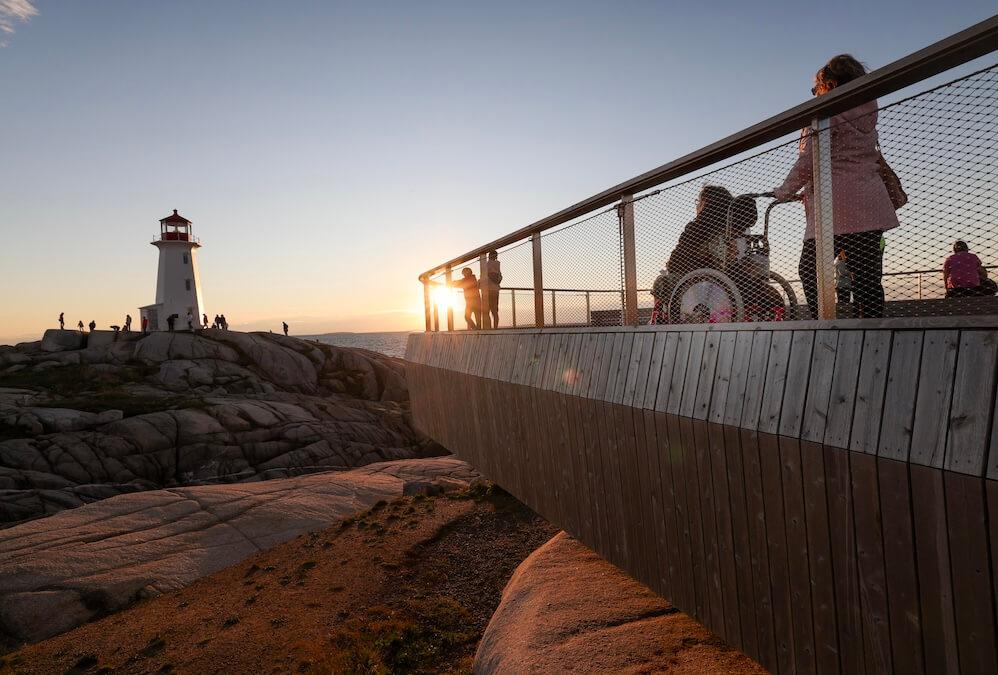People on boardwalk, including a person in a wheelchair, watching the sunset with the lighthouse in the background.
